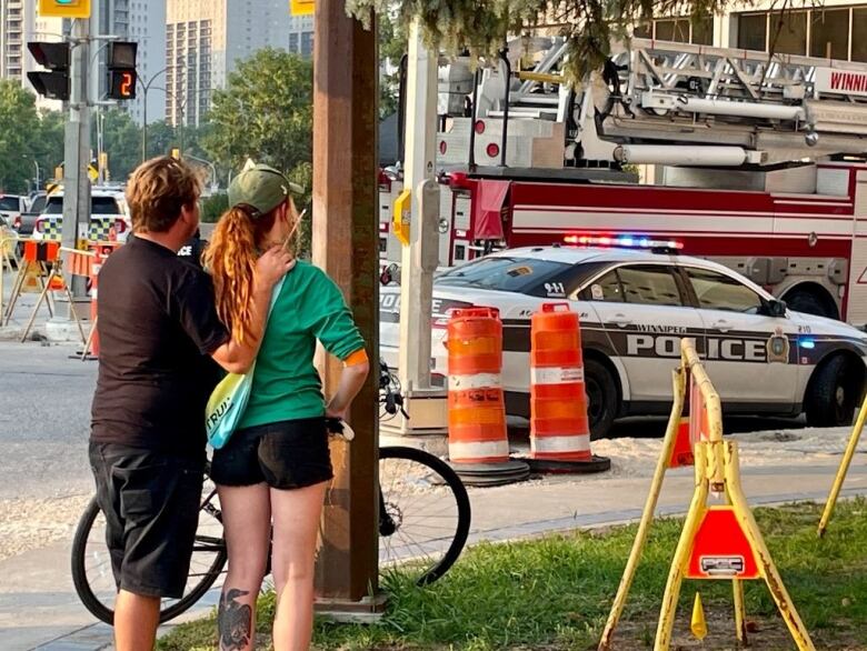 Two people stand near a traffic light near a police car and fire truck.