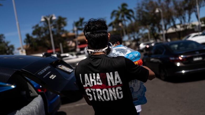 The back of a man wearing a t-shirt with the words Lahaina Strong is shown. The man is carrying a case of bottled water.