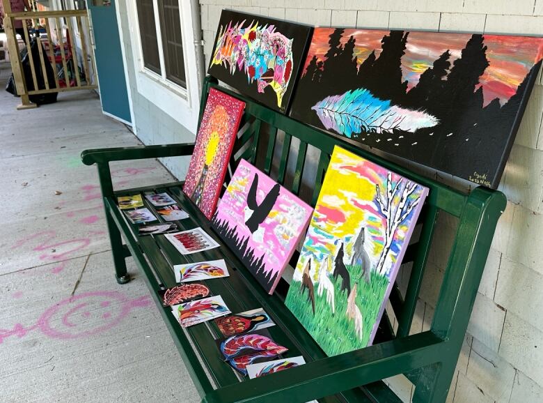 Multiple, colourful paintings sit on a green bench.