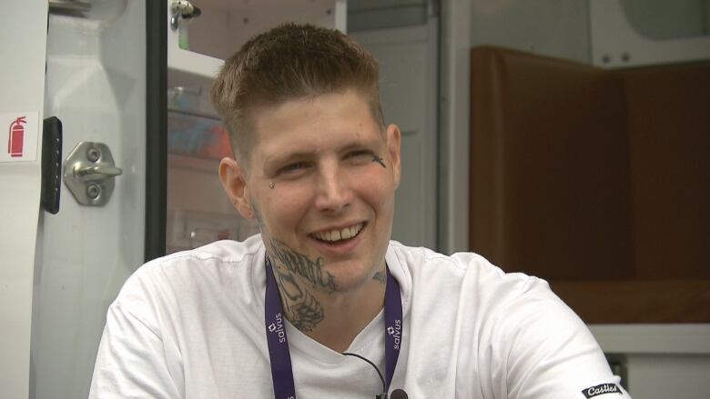 Young man with short brown hair and neck tattoos wearing short sleeved white T-shirt.