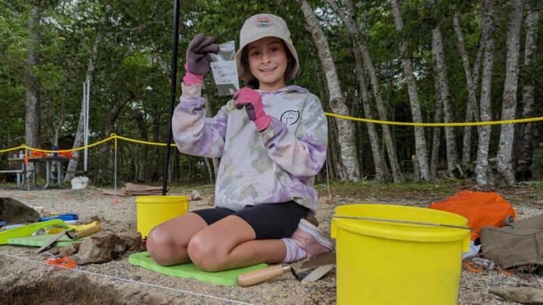 A little girl holds up a baggy with an artifact inside. She is smiling at the camera. She wears a white hat, purple sweater and pink gloves. 