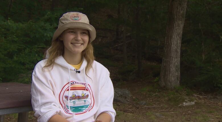 A girl with blonde hair smiles toward the camera. She is wearing a beige bucket hat and a white hoodie with a logo on it. 