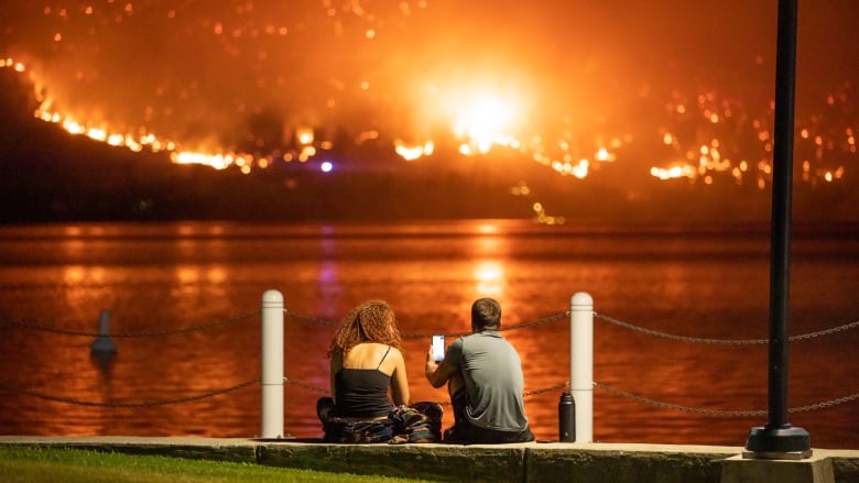 A young man and woman sit on the edge of a lake staring at wildfires on a hill across the water.