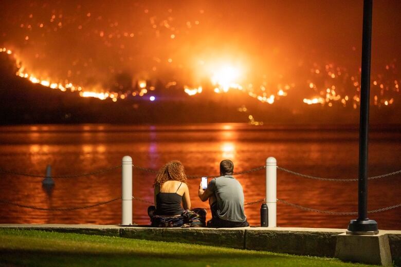 A young man and woman sit on the edge of a lake staring at wildfires on a hill across the water.