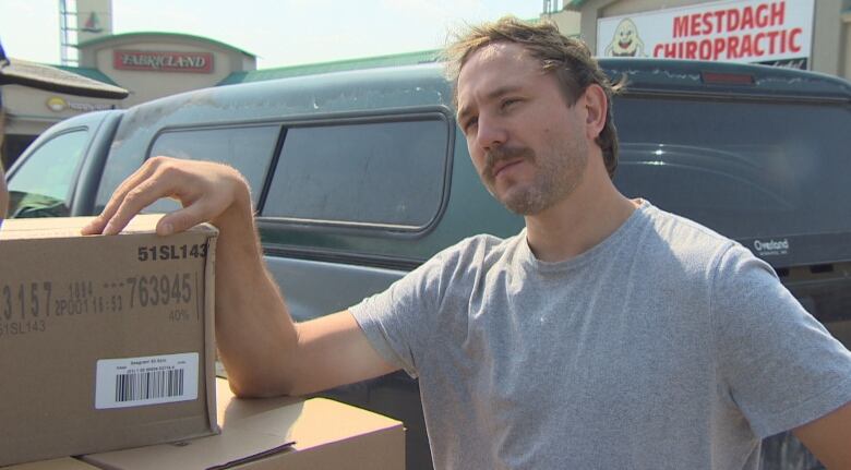 A man leans an arm on a box while looking at the camera. He has a brown moustache and short brown hair