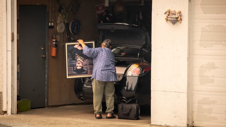 A woman with her back to the camera is packing belongings into a black sedan.