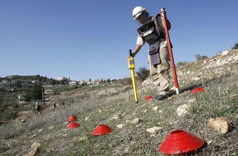 A woman in a long protective vest and white helmet, with a face shield, holds to long objects in her hands as she walks past various red markers in a rocky field. 