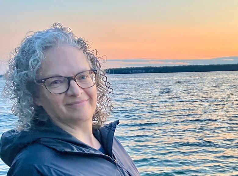 a woman wearing glasses, with curly hair, smiling in front of a water body