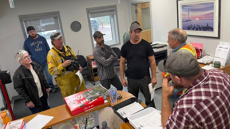 A group of men, some in work gear, stand in an office talking. 