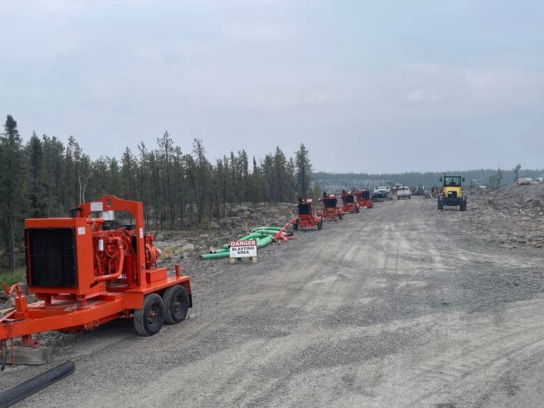 A line of industrial water pumps are seen lined up down a gravel road with construction trucks and equipment nearby.