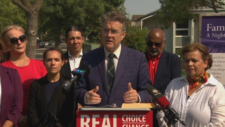 A man in a suit speaks at a podium surrounded by six people.