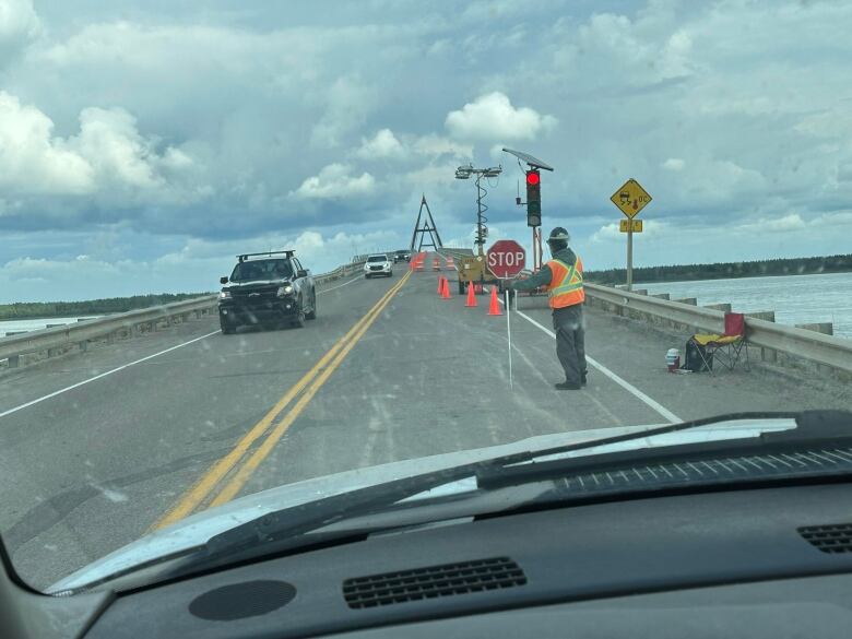 Vehicles are seen through a windshield crossing a bridge, with a highway worker holding a stop sign near some pylons on the road. 