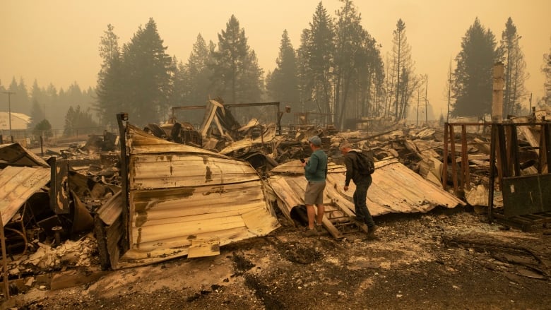 Two men wearing hiking boots stand looking at a huge pile of rubble and ash, with a smoky sky in the background.