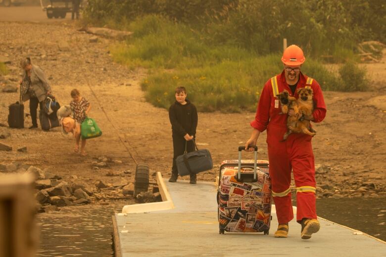 A man in a red jumpsuit carries two small brown puppies down onto a dock.