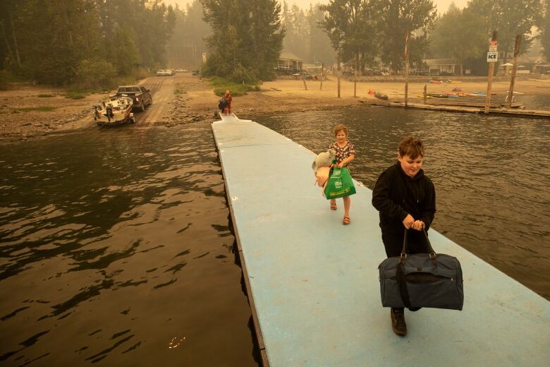 A boy and a girl carrying bags of clothing walk down a dock towards a boat.