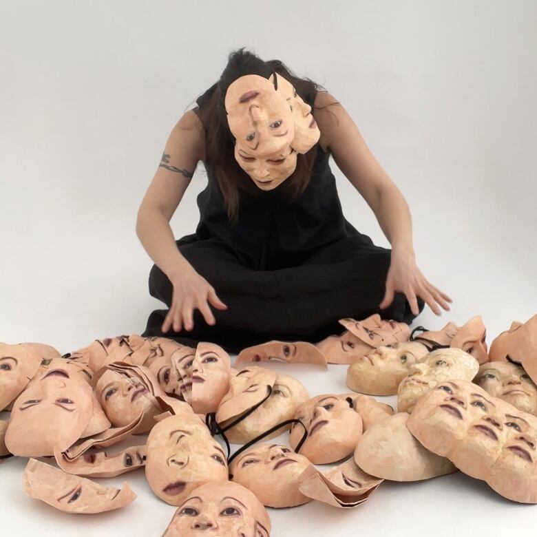 A woman wearing a realistic mask, seems to search among a pile of other human-like masks around her.