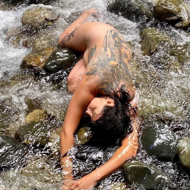 A nude woman with a large tattoo reclines restfully in a river.