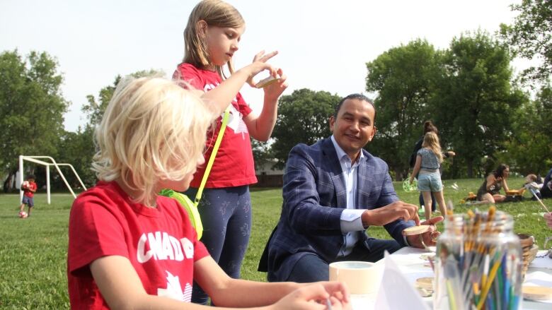 A man does some arts and crafts around a table, in a park, with some kids.