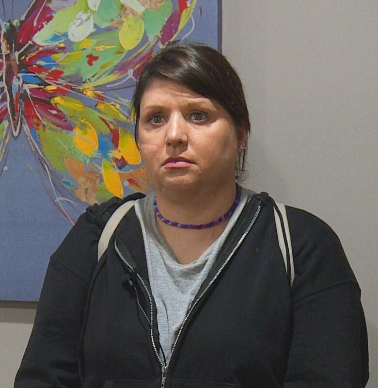 A woman wearing a black sweater over a grey shirt standing next to a painting of a butterfly