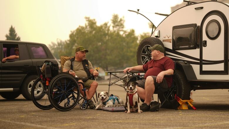 Two men in T-shirts and shorts sit on folding chairs beside a camping trailer against a hazy sky. Two small white and brown dogs sit between them.