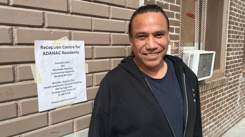 A man stands in front of a brown brick building. A signed that says 