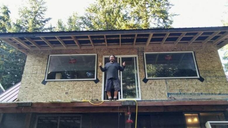 A man with long, wavy dark hair wearing shorts and work boots stands on the roof of a semi-finished two-storey home, giving a thumbs up.