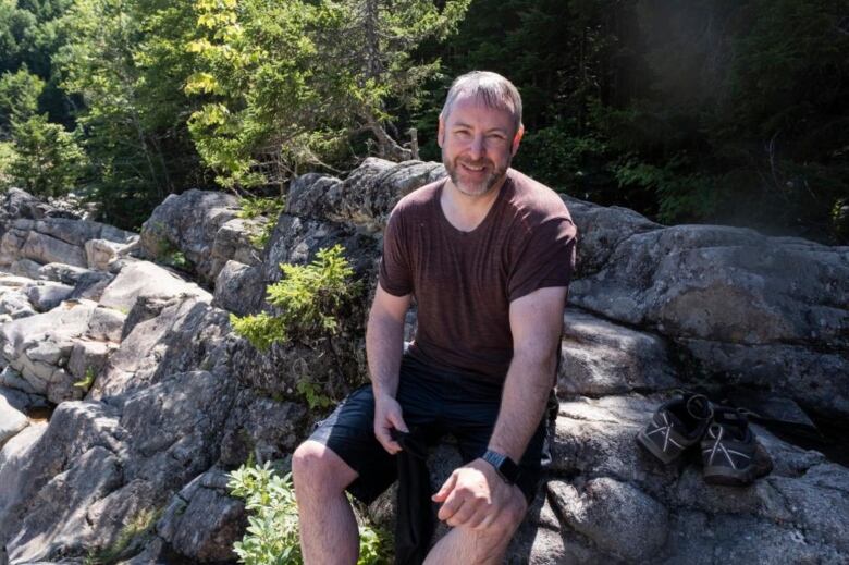 A man wearing a wet t-shirt sits on some rocks and smiles at the camera. His shoes are seen sitting on rocks to his right.