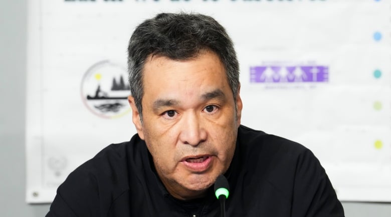 A First Nations chief speaks at a news conference. In the backdrop are the words 'What we do to the Earth we do to ourselves.'