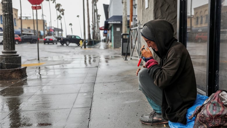 A person sits on a blue tarp on the sidewalk in the rain, head bowed and eyes closed. 