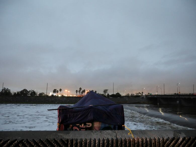 A tent on a pier perched right on the edge of the water. Palm trees, city lights and dark, cloudy skies are visible on the horizon.