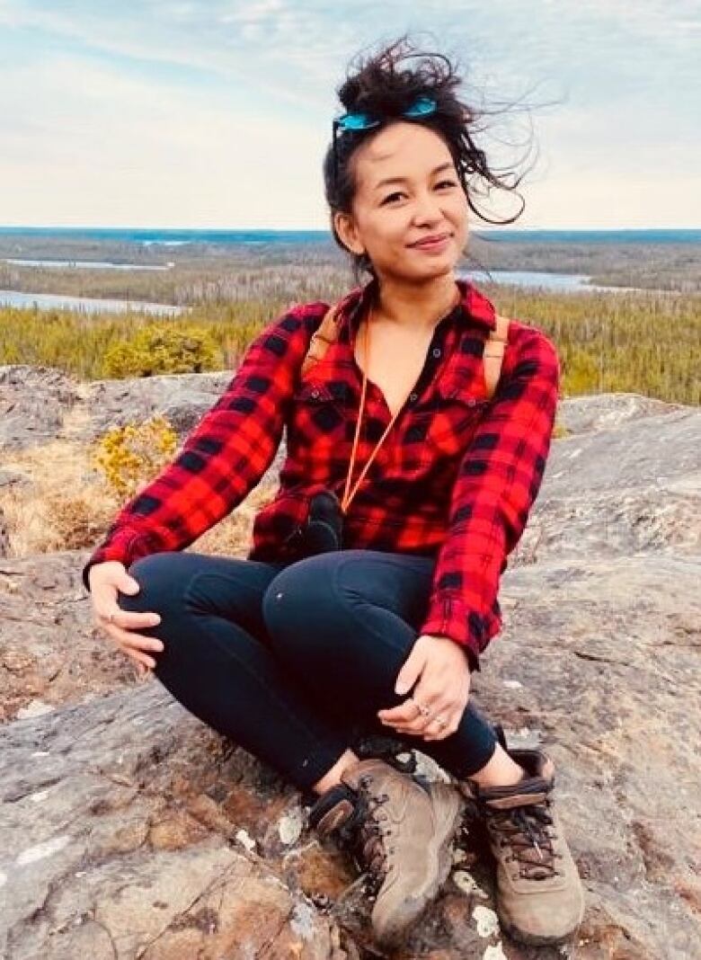 A woman wearing a red and black plaid shirt, dark blue jeans and brown hiking boots sits on a rock, smiling for a photo.