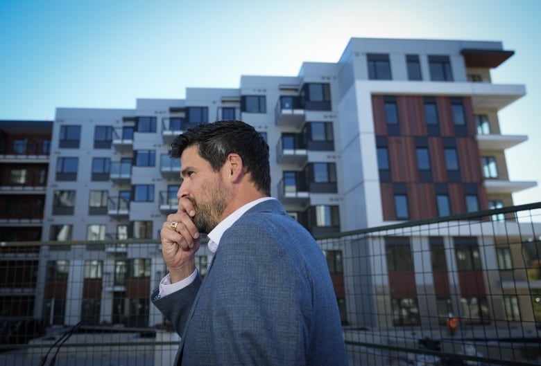 A politician stands to the side while an outdoor announcement in summer is happening. There's a condo-style building in the back.