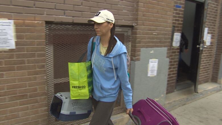 A woman in a baseball cap walks out of an apartment building carrying a suitcase, a cat carrier and a grocery bag.