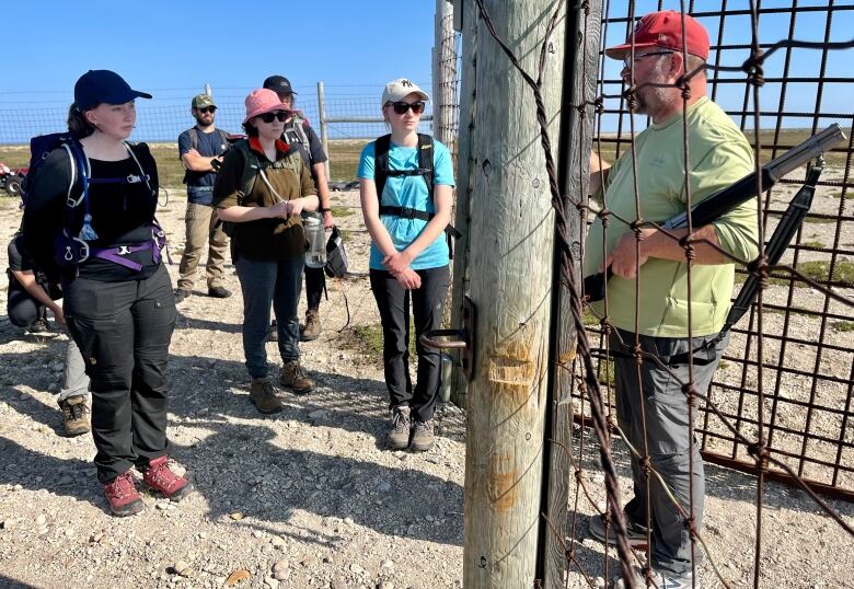 A man carrying a shotgun stands within a metal gate and speaks to students.