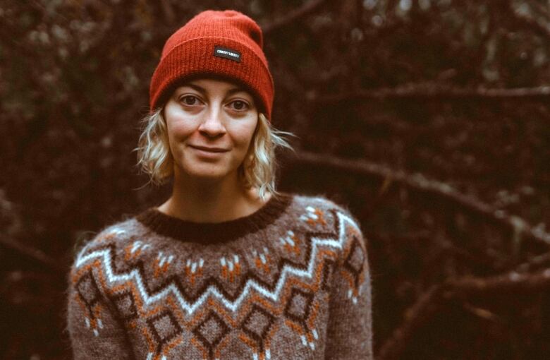 A woman in a grey sweater with a black, white and orange design stands in front of some trees.