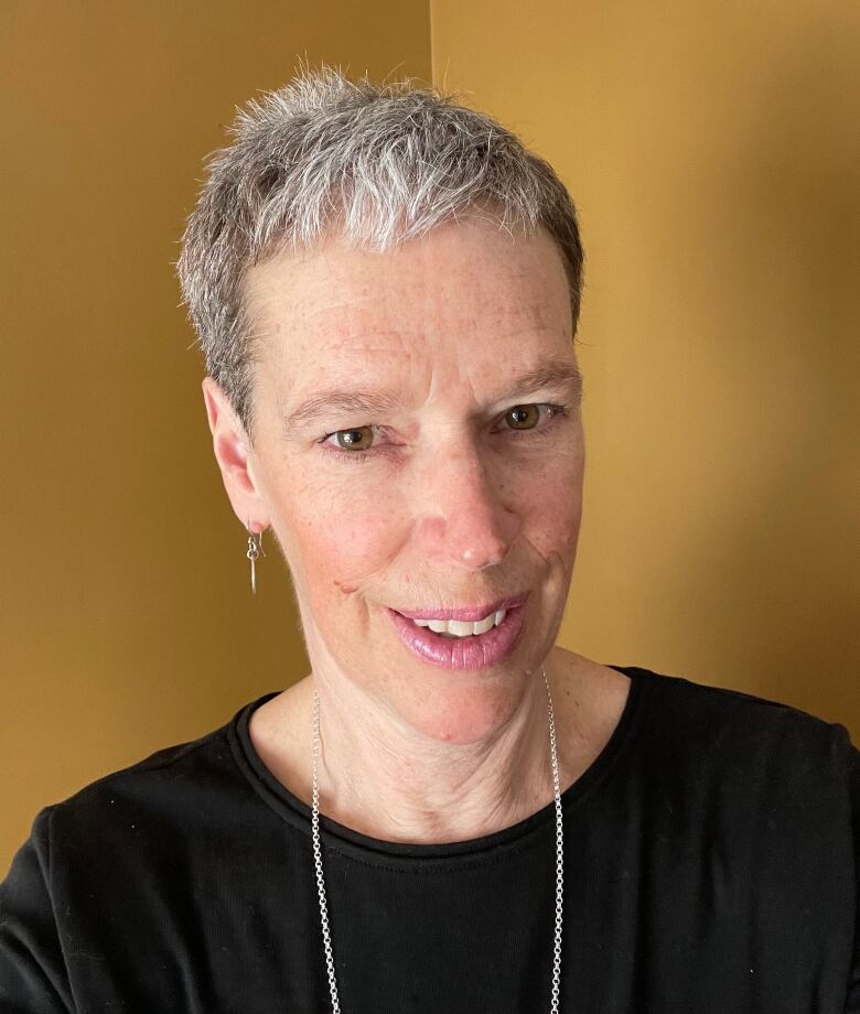 A woman with short silver hair smiles at the camera. She's wearing a black shirt, with yellow walls in the background.