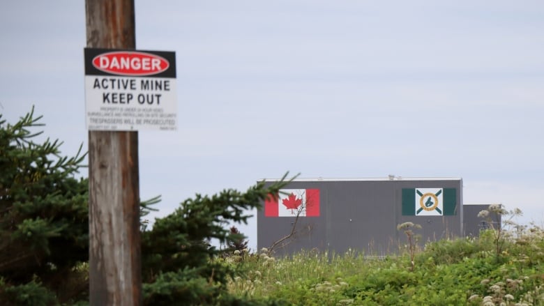 A large grey building with Canada and Cape Breton flags is shown in the distance, with a sign mounted on a pole in the foreground saying 'Danger Active Mine Keep Out.'