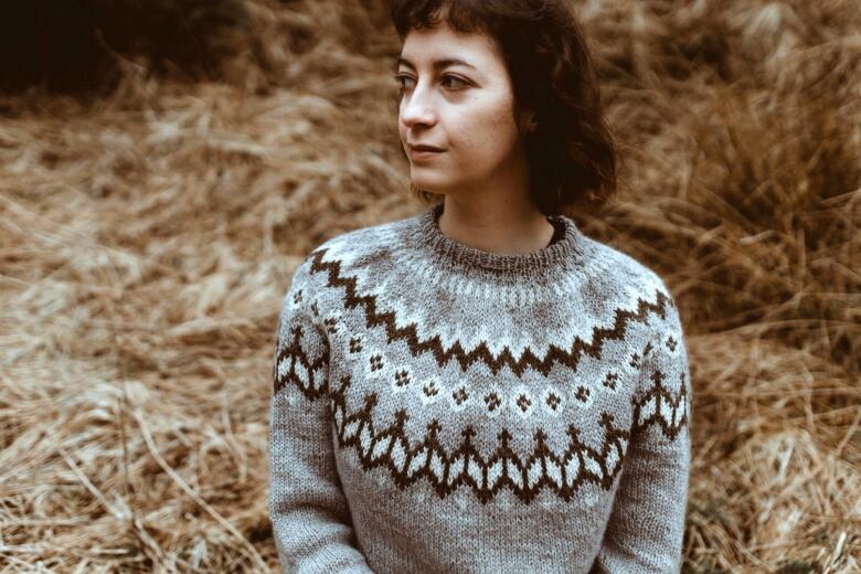 A woman with dark hair sits in a hay-covered field, wearing a grey sweater with a brown and white pattern.