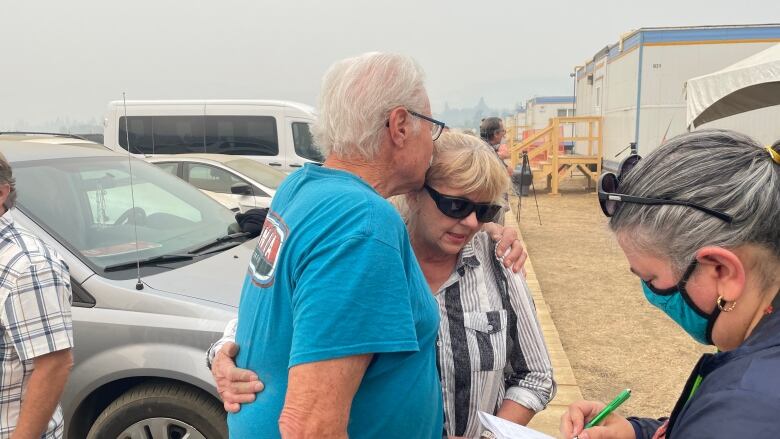 A man with white hair in a blue T-shirt kisses a woman in a blouse and dark sunglasses.