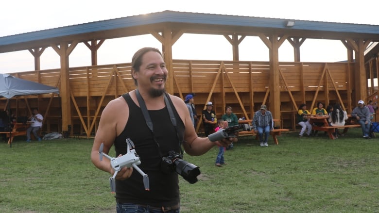 A First Nations man holding camera gear smiles and walks in a powwow arbor.