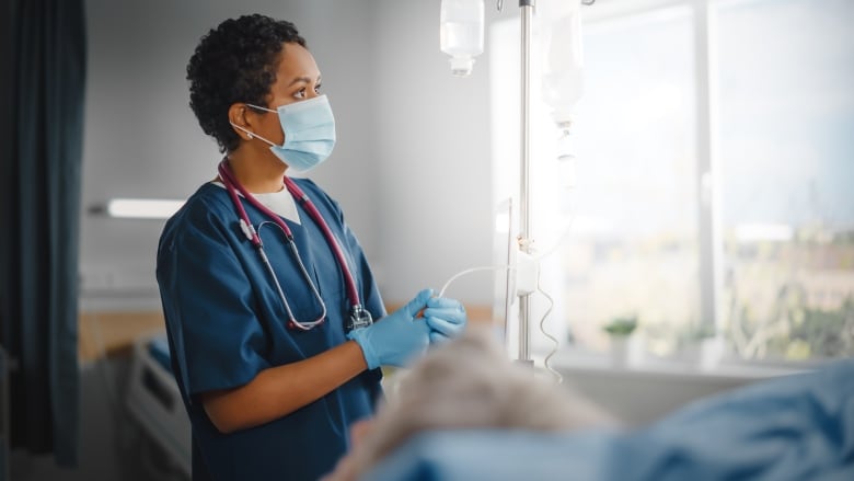 A woman in a surgical mask wearing blue hospital gloves and dark blue medical garb stands by a patient's bed beside a window with an outside view, taking vitals.