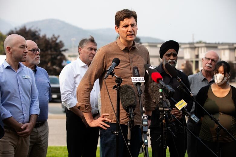 A tall white man wearing a brown shirt speaks at an outdoor news conference, flanked by numerous others.