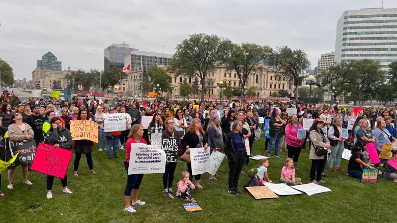 A large group of people attending a rally are seen standing on a stretch of grass, with some holding signs.