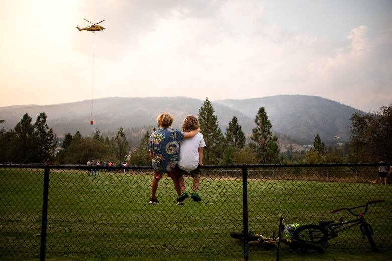 Two children sitting on a wire fence watching a helicopter, with smokey from wildfire and mountain range in the background.