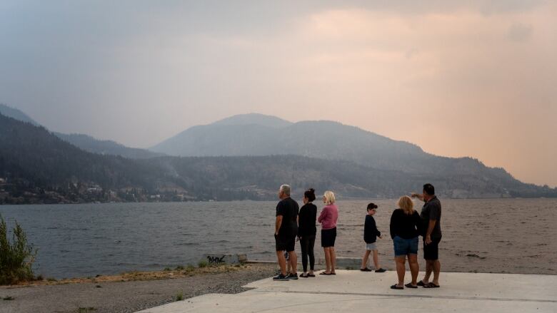 A group of six people stand next to a lake and look across at a hillside shrouded in light smoke and clouds.