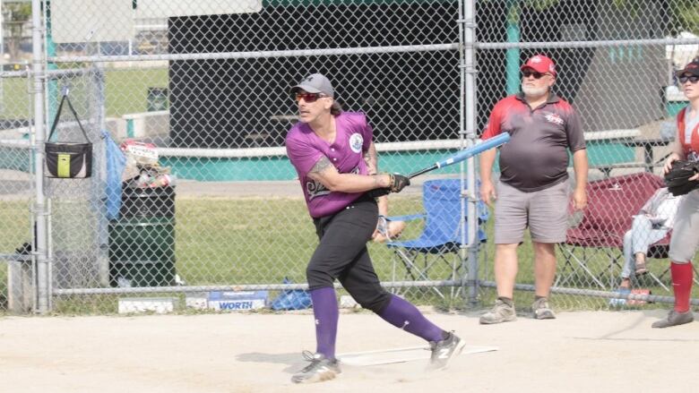 A person in a purple baseball jersey and purple socks follows through a swing of the bat and gets ready to run.