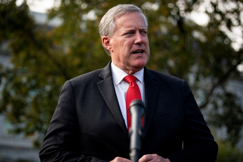 Grey haored man wearing a dark suit and red tie speaks into a microphone