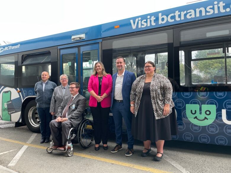 Six people stand in front of a bus. The third one from the left is in a wheelchair.