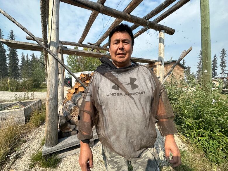 A man in a bug net standing in front of a wooden framed structure.