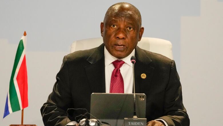 A man seated in a suit is shown speaking into a microphone, with a small flag displayed nearby.